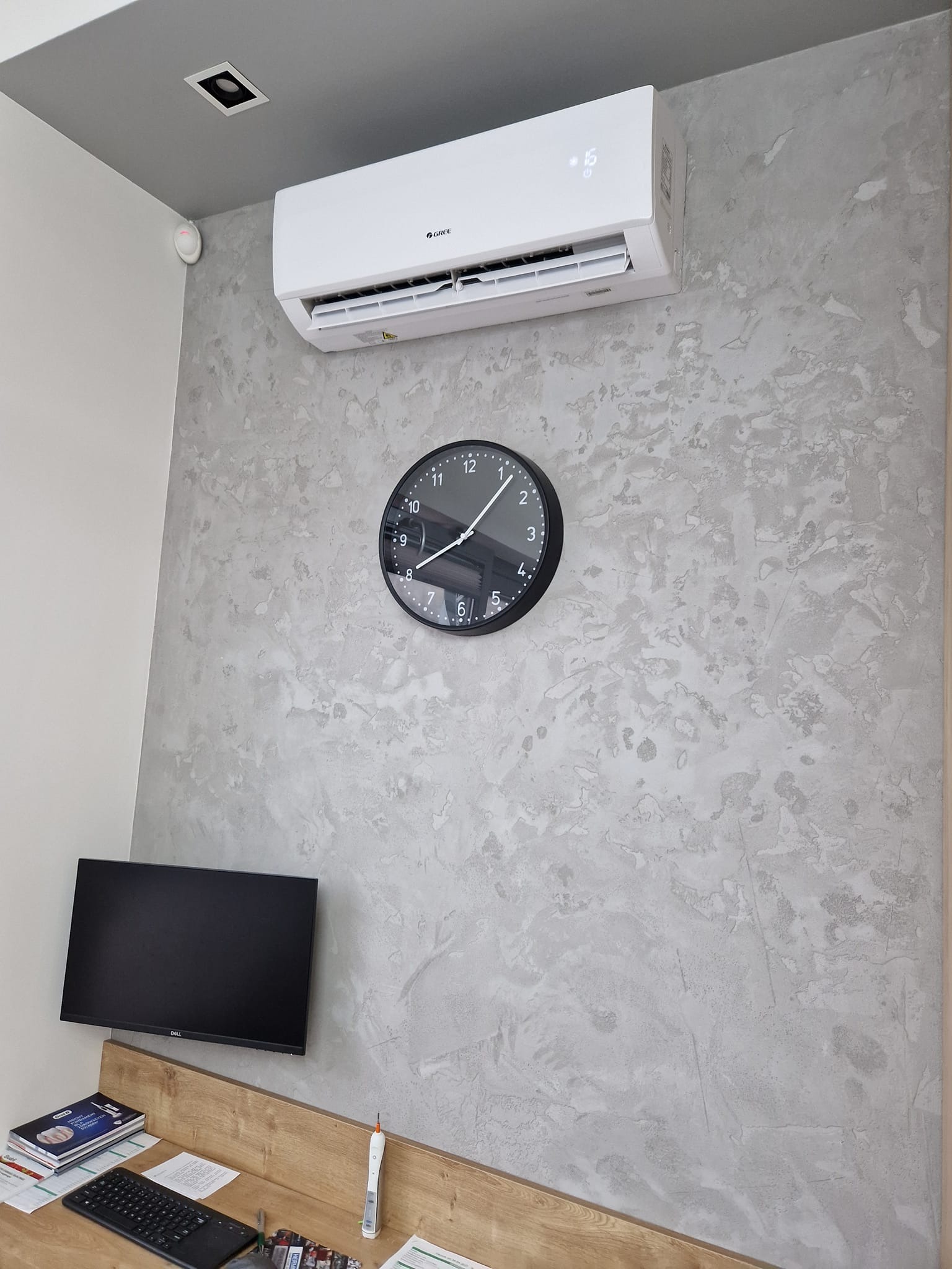 Biała jednostka klimatyzacyjna marki Gree zamontowana na dekoracyjnej, szarej ścianie obok czarnego zegara ściennego. Pod spodem widoczny biurkowy monitor Dell i klawiatura.