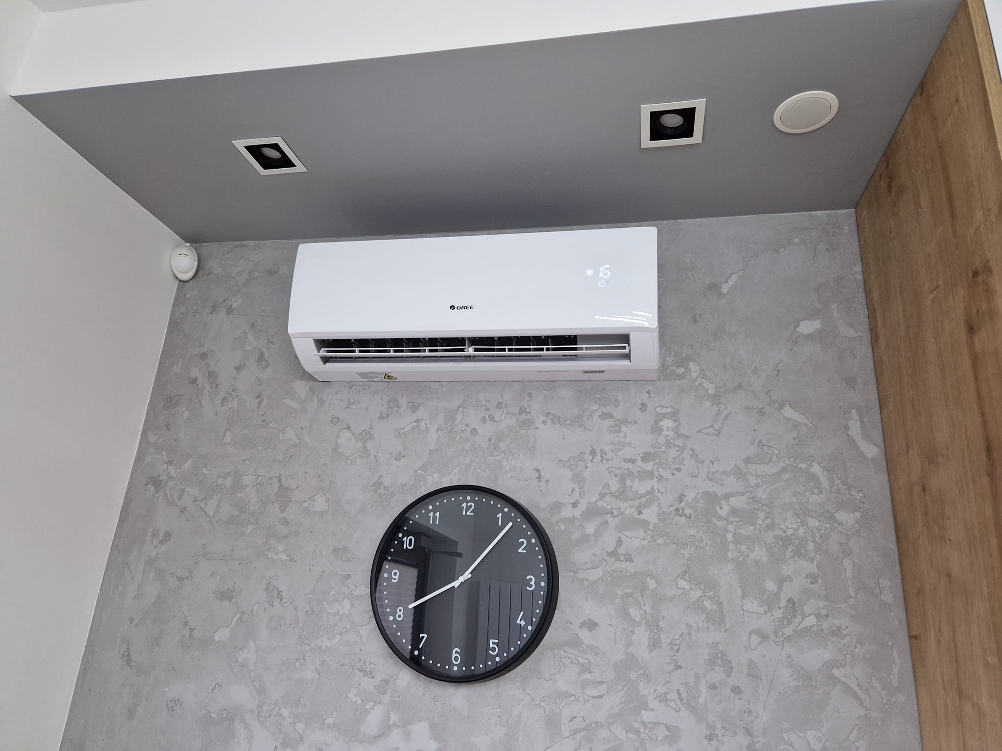 Biały klimatyzator marki Gree zamontowany na ścianie o szarej, teksturowanej powierzchni, pod nim czarny zegar wskazujący godzinę 11:10, w tle fragment drewnianej boazerii i szary sufit...
