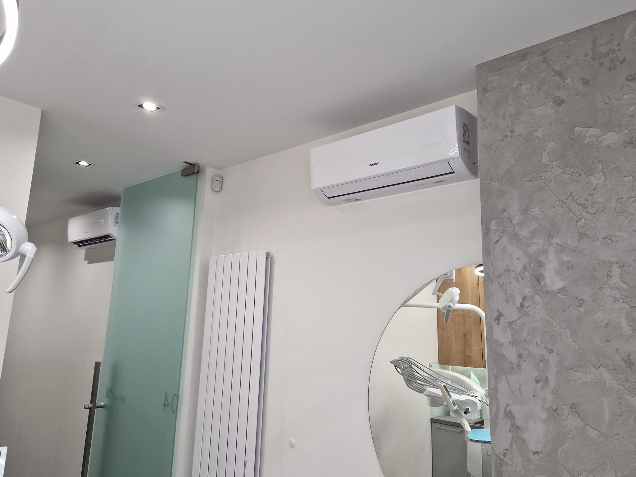 Biała klimatyzacja Gree zamontowana w gabinecie dentystycznym z widocznym fotelem i lampą dentystyczną w lustrze, obok dekoracyjna ściana w odcieniach szarości.
