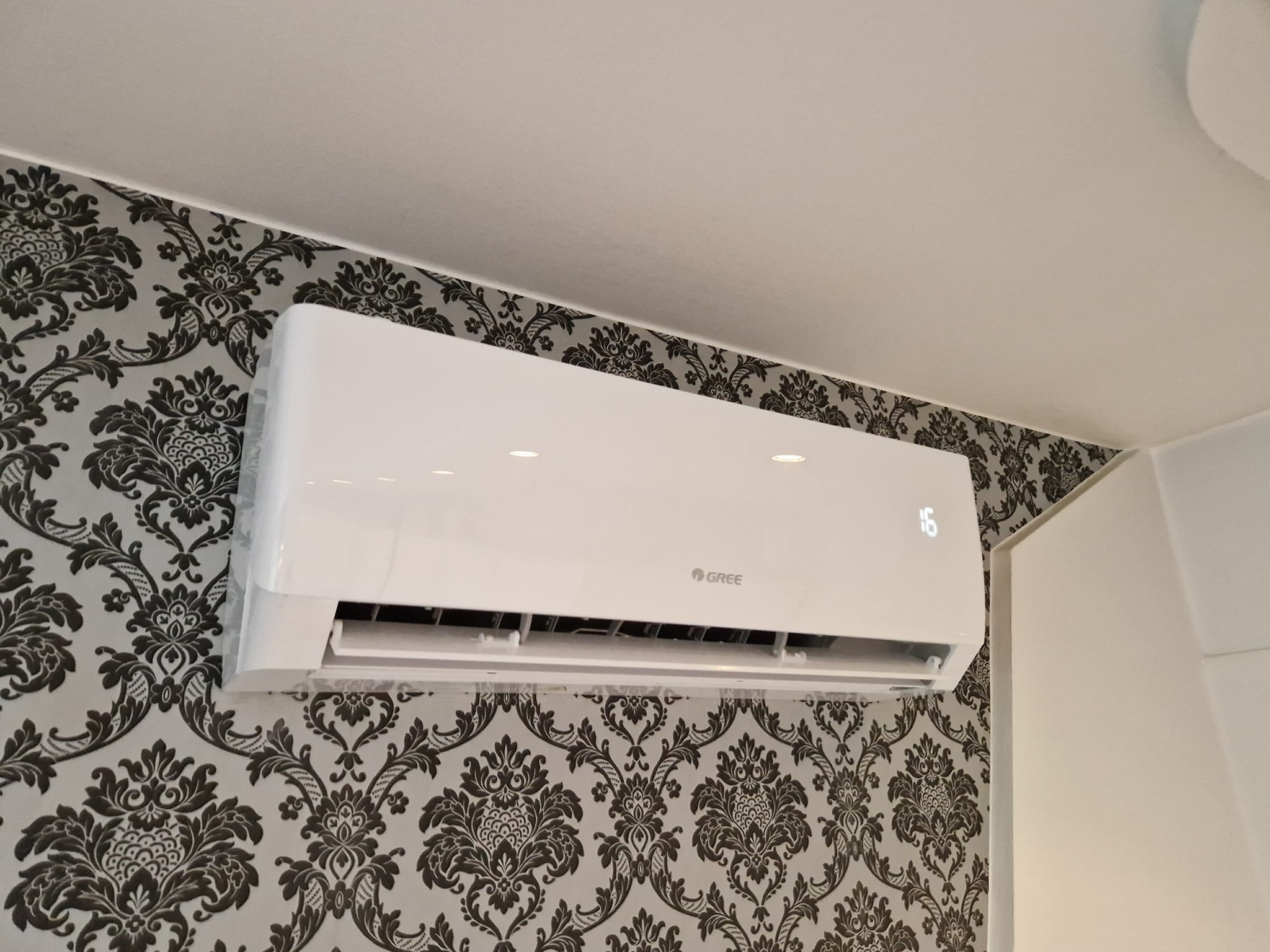 Biała jednostka klimatyzacji marki Gree zamontowana na ścianie z ozdobną tapetą w kwiatowe wzory, wyświetlacz pokazuje temperaturę 16 stopni.