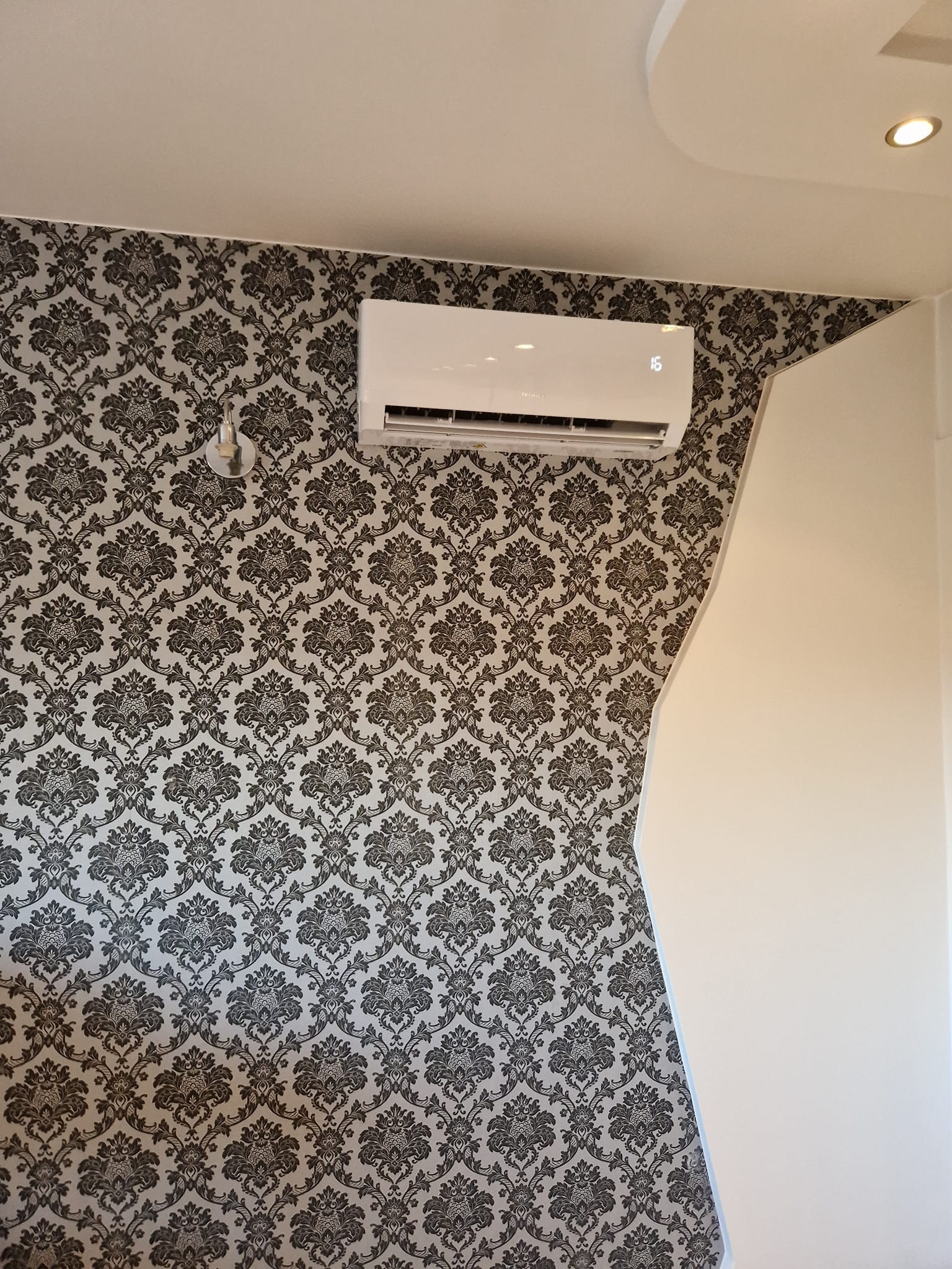 Biały klimatyzator ścienny zamontowany na ścianie z wzorzystą tapetą w stylu vintage, widoczny fragment białej ściany obok.