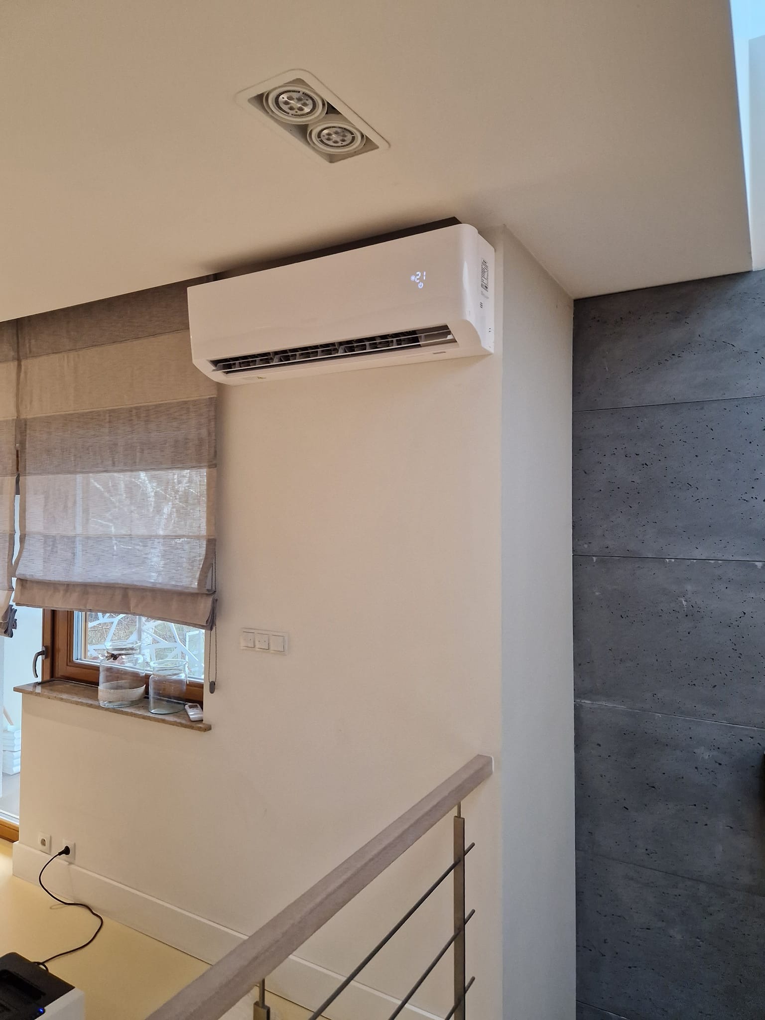 Biały klimatyzator ścienny zamontowany w nowoczesnym wnętrzu z szarymi roletami rzymskimi i ścianą z szarego kamienia dekoracyjnego.