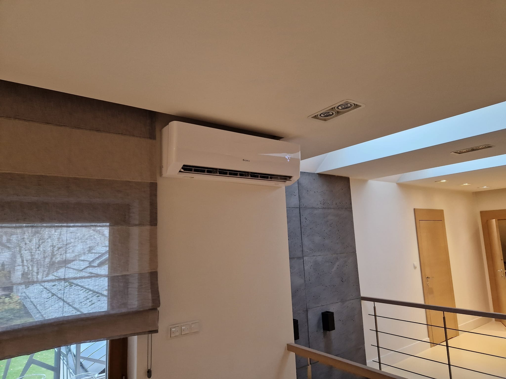 Biały klimatyzator marki Gree zamontowany na ścianie w nowoczesnym wnętrzu, widoczne rolety rzymskie, szara ściana dekoracyjna i balustrada.