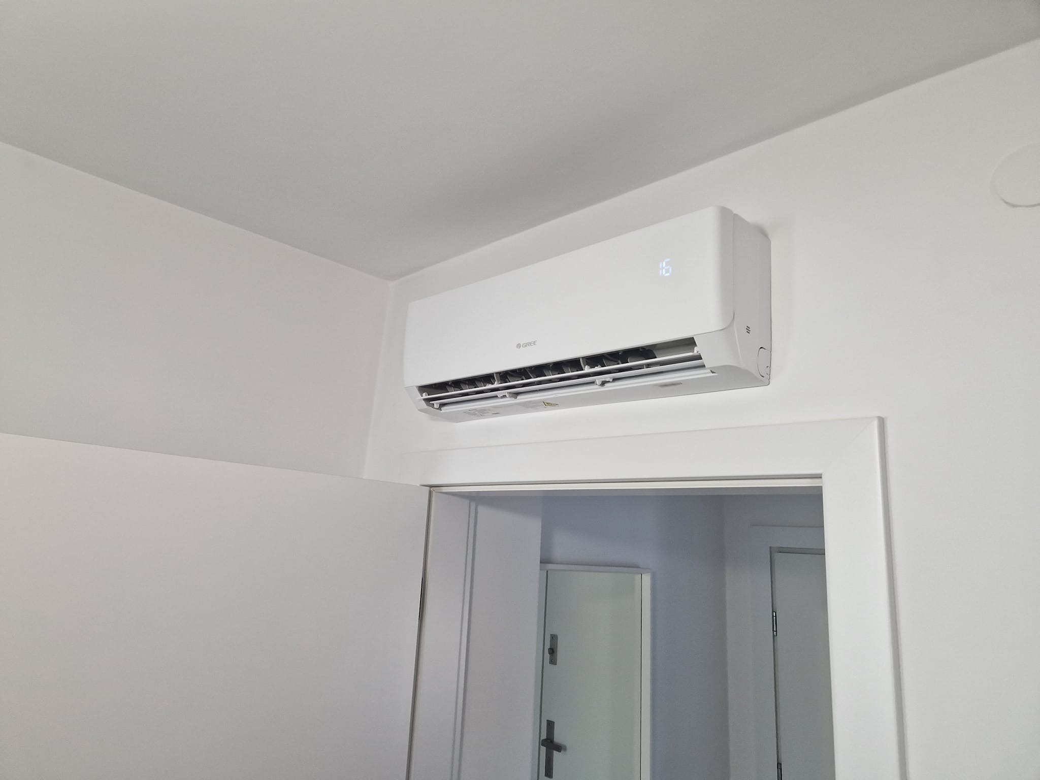 Biały klimatyzator marki Gree zamontowany na ścianie w pokoju, widoczny wyświetlacz temperatury, drzwi w tle.