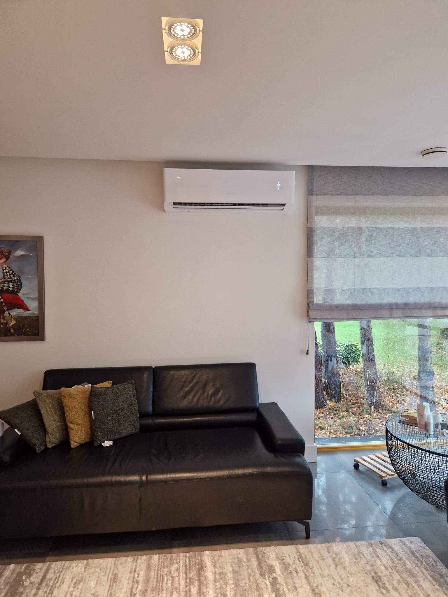 Biały klimatyzator ścienny zamontowany w salonie nad czarną skórzaną sofą, widok na zewnątrz przez okno z szarymi roletami rzymskimi.