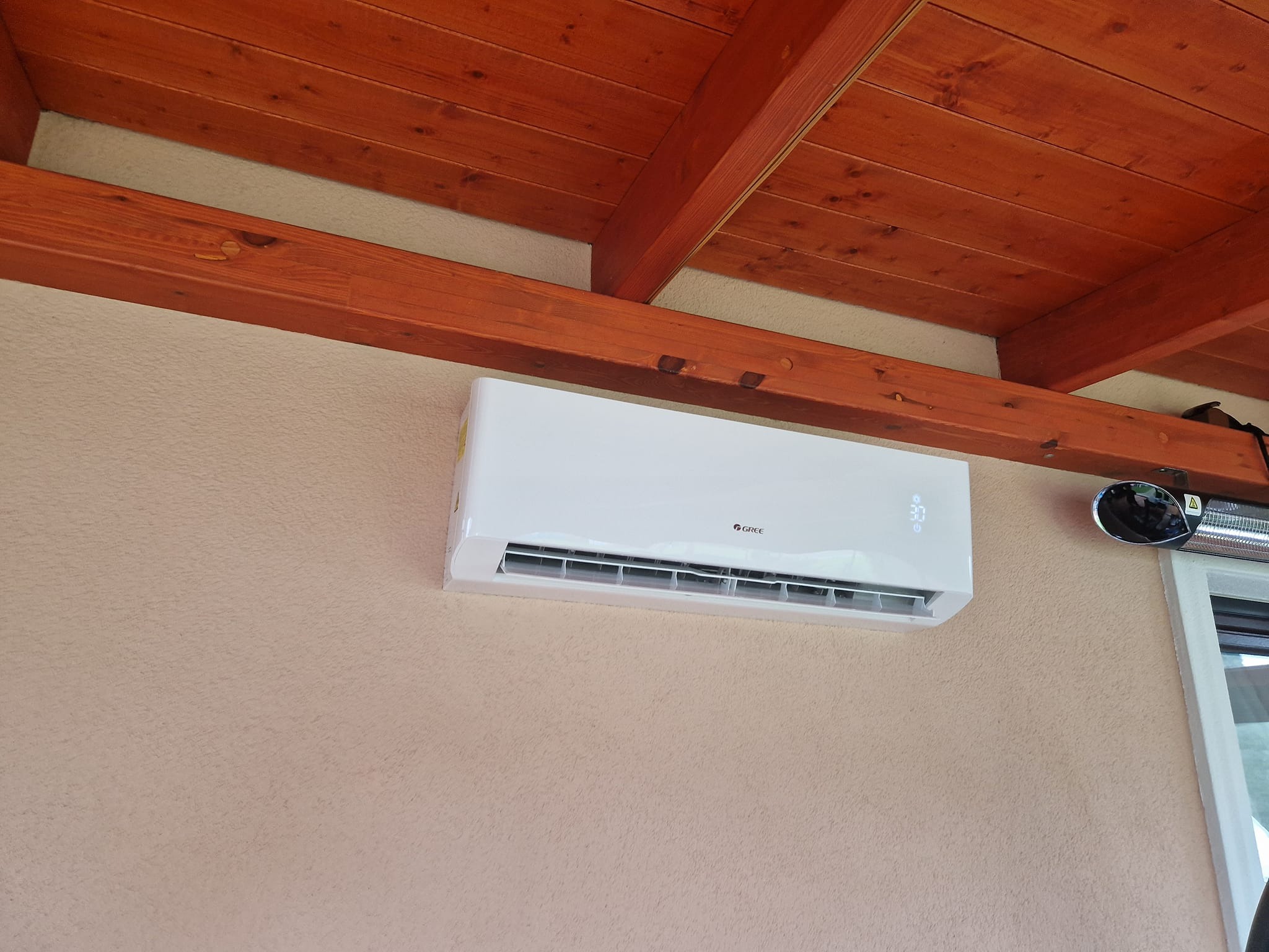 Biały klimatyzator Gree zamontowany na teksturowanej ścianie wewnątrz pomieszczenia z drewnianym sufitem i widocznym grzejnikiem zewnętrznym.