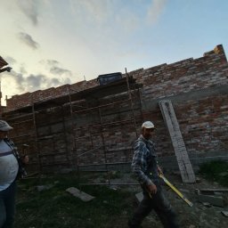 Firma Ogólnobudowlana Chwastyniak - Dwóch murarzy przy wznoszeniu ściany z czerwonej cegły, widoczne rusztowanie, jeden z pracowników trzyma poziomicę, drugi młotek.