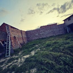 Firma Ogólnobudowlana Chwastyniak - Wznoszenie muru z czerwonej cegły na nowo budowanym domu, widoczna drabina oparta o ścianę, trawa i pochmurne niebo w tle.