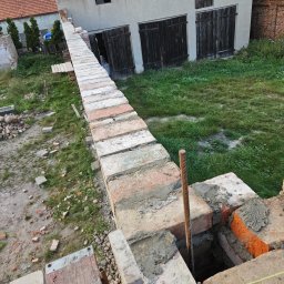 Firma Ogólnobudowlana Chwastyniak - Wznoszenie muru z czerwonej cegły na zaprawie cementowej, widoczne elementy zbrojenia pionowego, w tle budynek gospodarczy z drewnianymi wrotami i zarośnięty teren.