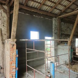 Firma Ogólnobudowlana Chwastyniak - Wnętrze budynku gospodarczego w trakcie remontu, widoczne drewniane elementy konstrukcyjne, fragment ściany po tynkowaniu, metalowe bariery i resztki materiałów budowlanych na podłodze.