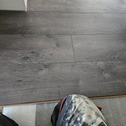 Firma Ogólnobudowlana Chwastyniak - Ułożone panele podłogowe w odcieniach szarości z widocznymi słojami drewna, przycięte przy ścianie, w tle fragment nogawki spodni roboczych ubrudzonych farbą.