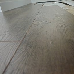 Firma Ogólnobudowlana Chwastyniak - Układanie paneli podłogowych z widoczną strukturą drewna, fragment podkładu i listwa przypodłogowa w tle.