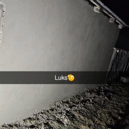 Firma Ogólnobudowlana Chwastyniak - Świeżo otynkowana ściana domu z widoczną strukturą tynku, oświetlona od dołu, z ziemią i fragmentem dachu w kadrze. Na zdjęciu widoczny napis 'Luks' z emotikoną.