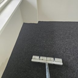 Firma Ogólnobudowlana Chwastyniak - Wałek do tapetowania na ciemnoszarym, ziarnistym tle, prawdopodobnie ściany, blisko białej ściany i okna.