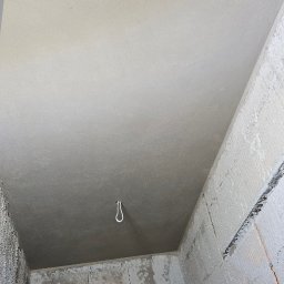 Firma Ogólnobudowlana Chwastyniak - Wnętrze w trakcie prac wykończeniowych: gładki, jasnoszary sufit po szpachlowaniu, surowe ściany z betonu komórkowego z widocznymi śladami zaprawy, wystający kabel oświetleniowy, narzędzia...