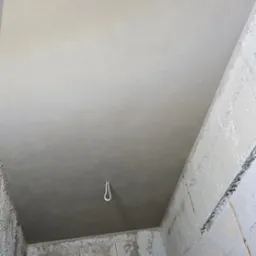Wnętrze w trakcie prac wykończeniowych: gładki, jasnoszary sufit po szpachlowaniu, surowe ściany z betonu komórkowego z widocznymi śladami zaprawy, wystający kabel oświetleniowy, narzędzia...
