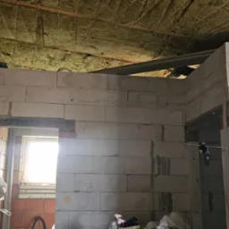 Wnętrze surowego domu w trakcie budowy: ściany z białych bloczków betonowych, częściowo otynkowane, z widocznymi przewodami elektrycznymi, izolacja z wełny mineralnej na suficie, surowa podłoga...