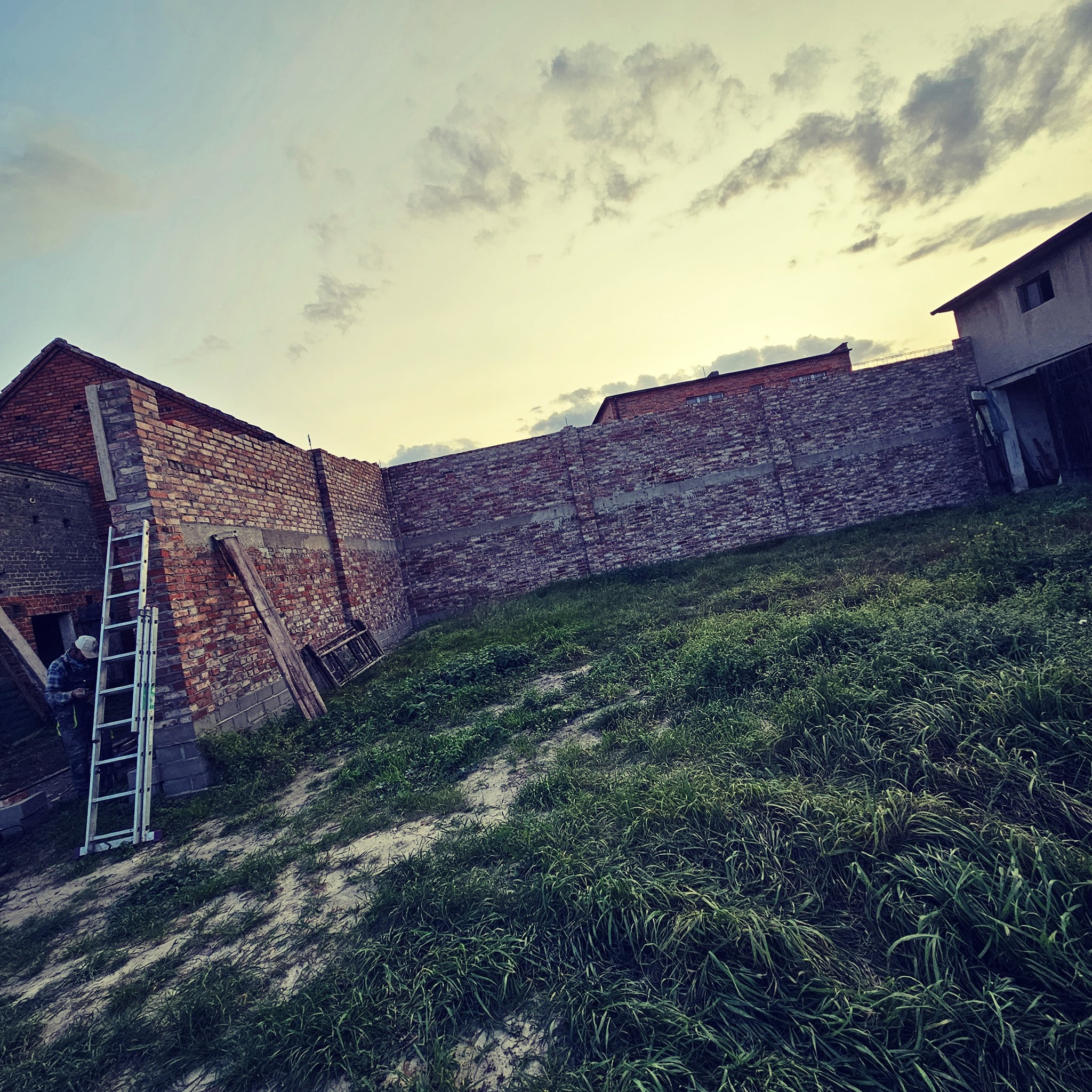 Wznoszenie muru z czerwonej cegły na nowo budowanym domu, widoczna drabina oparta o ścianę, trawa i pochmurne niebo w tle.