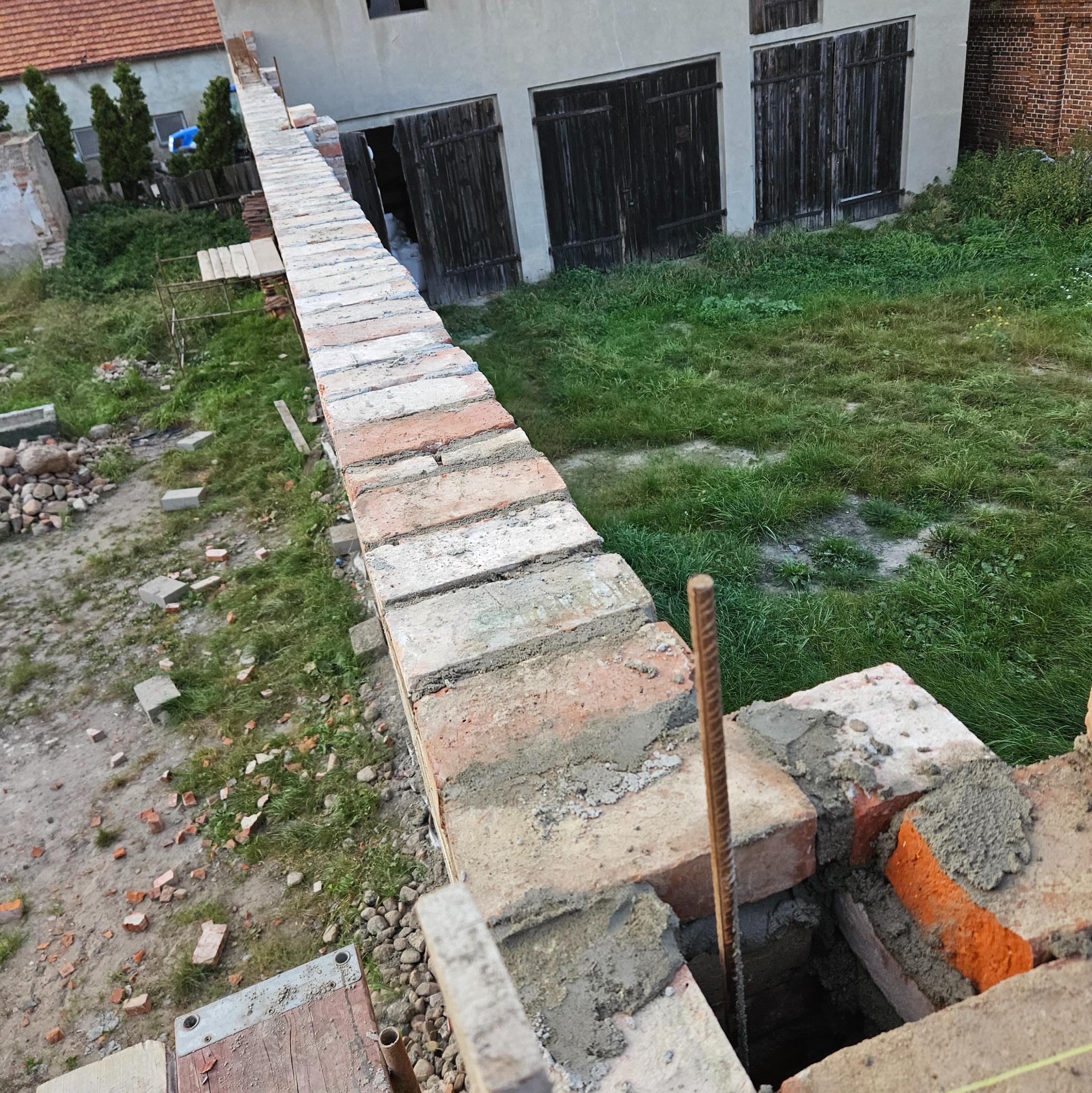 Wznoszenie muru z czerwonej cegły na zaprawie cementowej, widoczne elementy zbrojenia pionowego, w tle budynek gospodarczy z drewnianymi wrotami i zarośnięty teren.