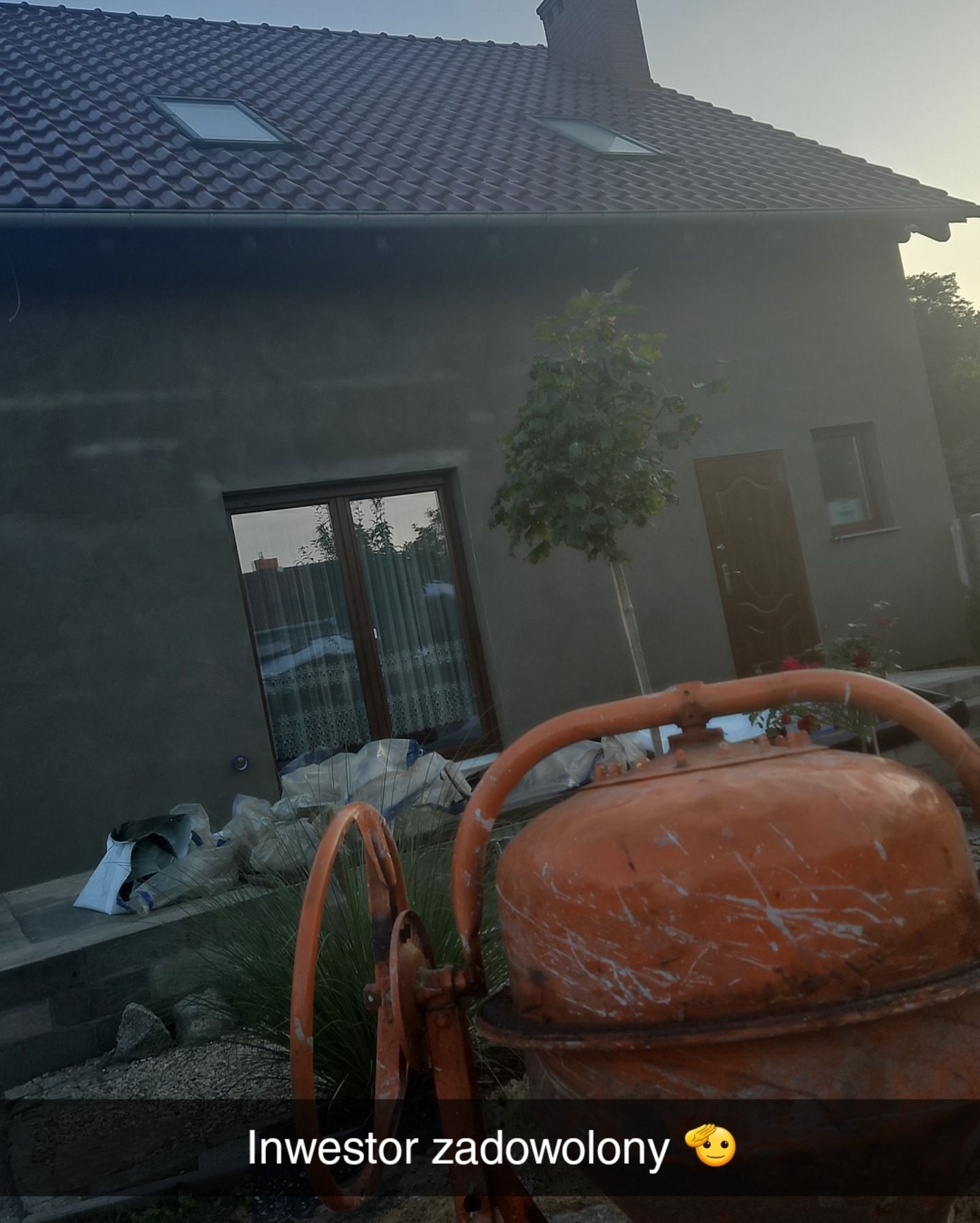 Surowy dom z szarą elewacją, widoczna dachówka i okno dachowe, na pierwszym planie brudna betoniarka i worki z materiałami budowlanymi.