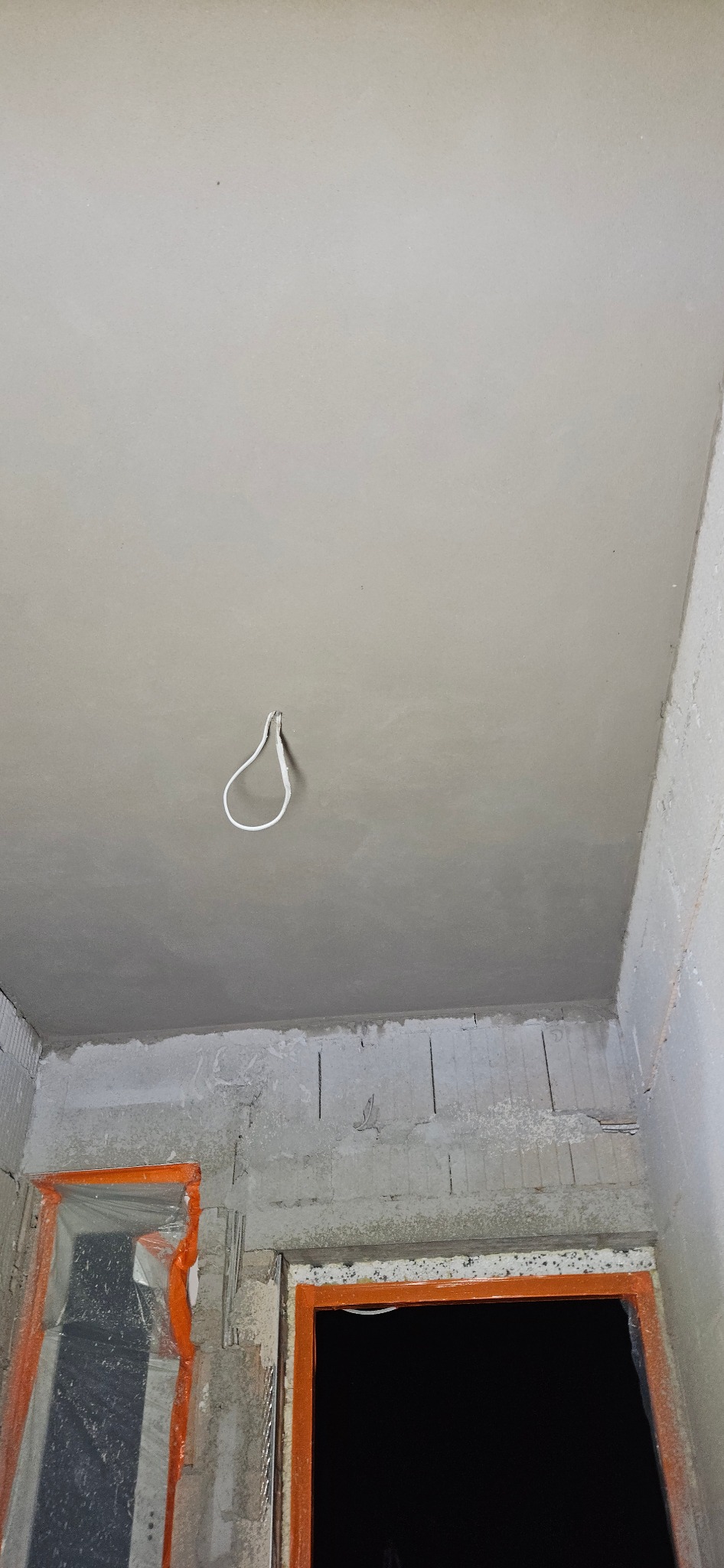 Gładki, szary sufit po nałożeniu gładzi szpachlowej, z wiszącym przewodem elektrycznym, widok na surowe ściany z zabezpieczonymi otworami okiennymi.