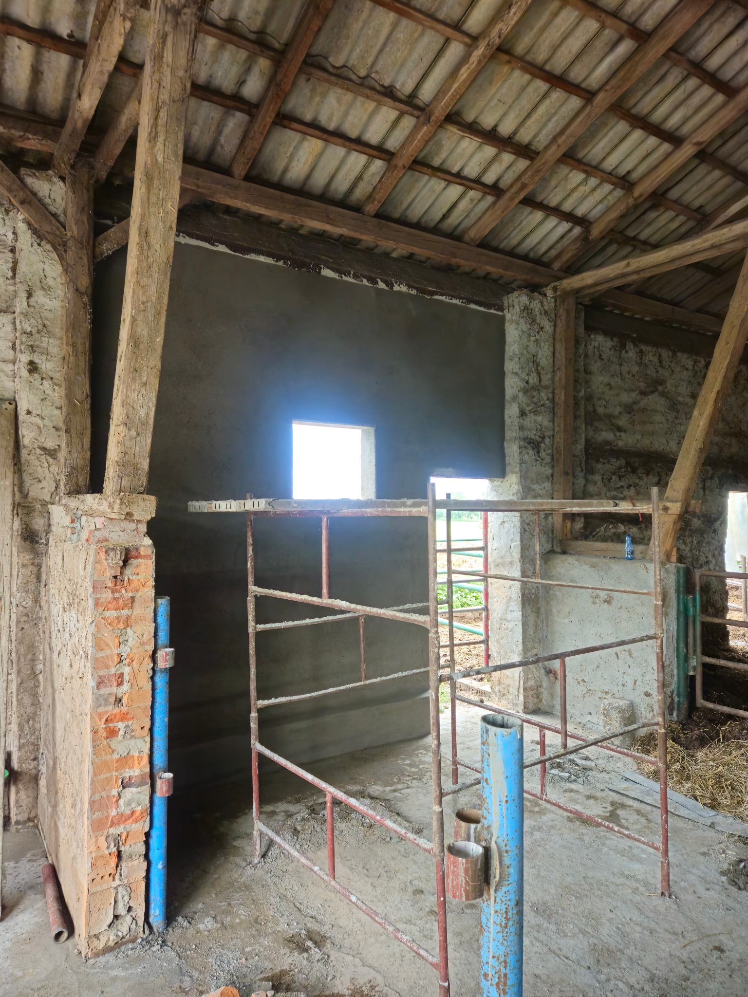 Wnętrze budynku gospodarczego w trakcie remontu, widoczne drewniane elementy konstrukcyjne, fragment ściany po tynkowaniu, metalowe bariery i resztki materiałów budowlanych na podłodze.