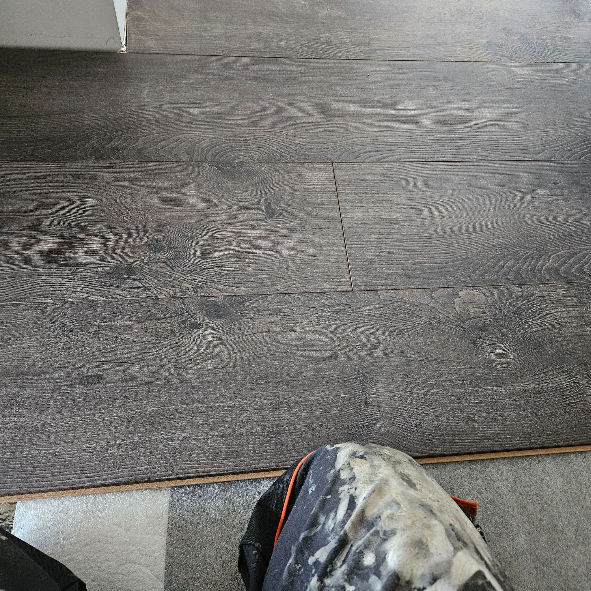 Ułożone panele podłogowe w odcieniach szarości z widocznymi słojami drewna, przycięte przy ścianie, w tle fragment nogawki spodni roboczych ubrudzonych farbą.