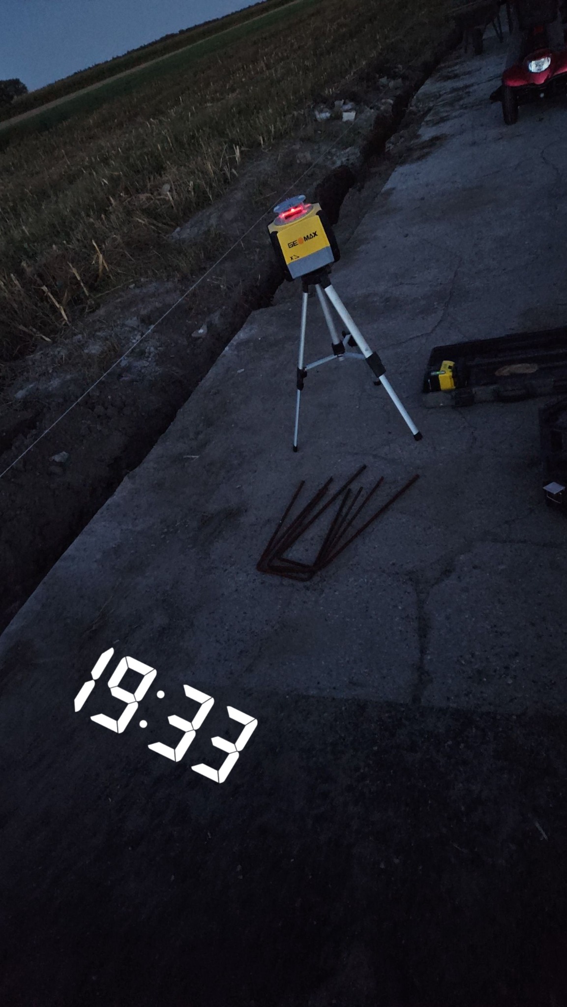 Żółty laser krzyżowy GeoMax na trójnogu ustawiony obok wykopu i prętów zbrojeniowych podczas prac ziemnych o zmierzchu.