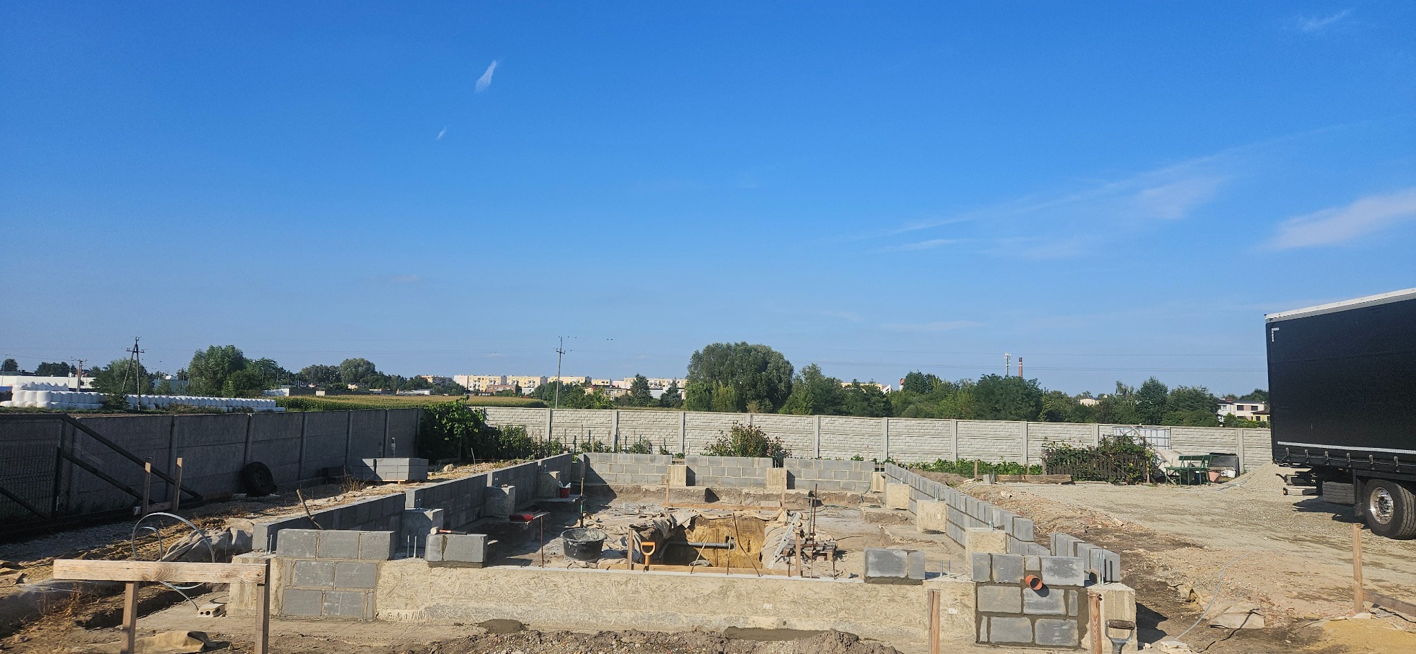 Budowa fundamentów domu z bloczków betonowych na działce z widokiem na odległe budynki mieszkalne, przy częściowo zaparkowanej ciężarówce