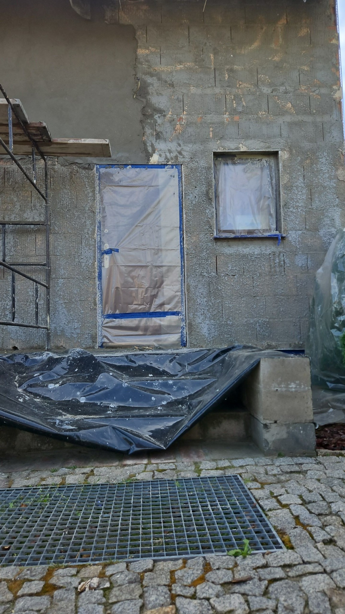 Fragment ściany budynku w trakcie prac budowlanych: część ściany otynkowana, okno i drzwi zabezpieczone folią, widoczne rusztowanie i czarna folia ochronna na schodach.