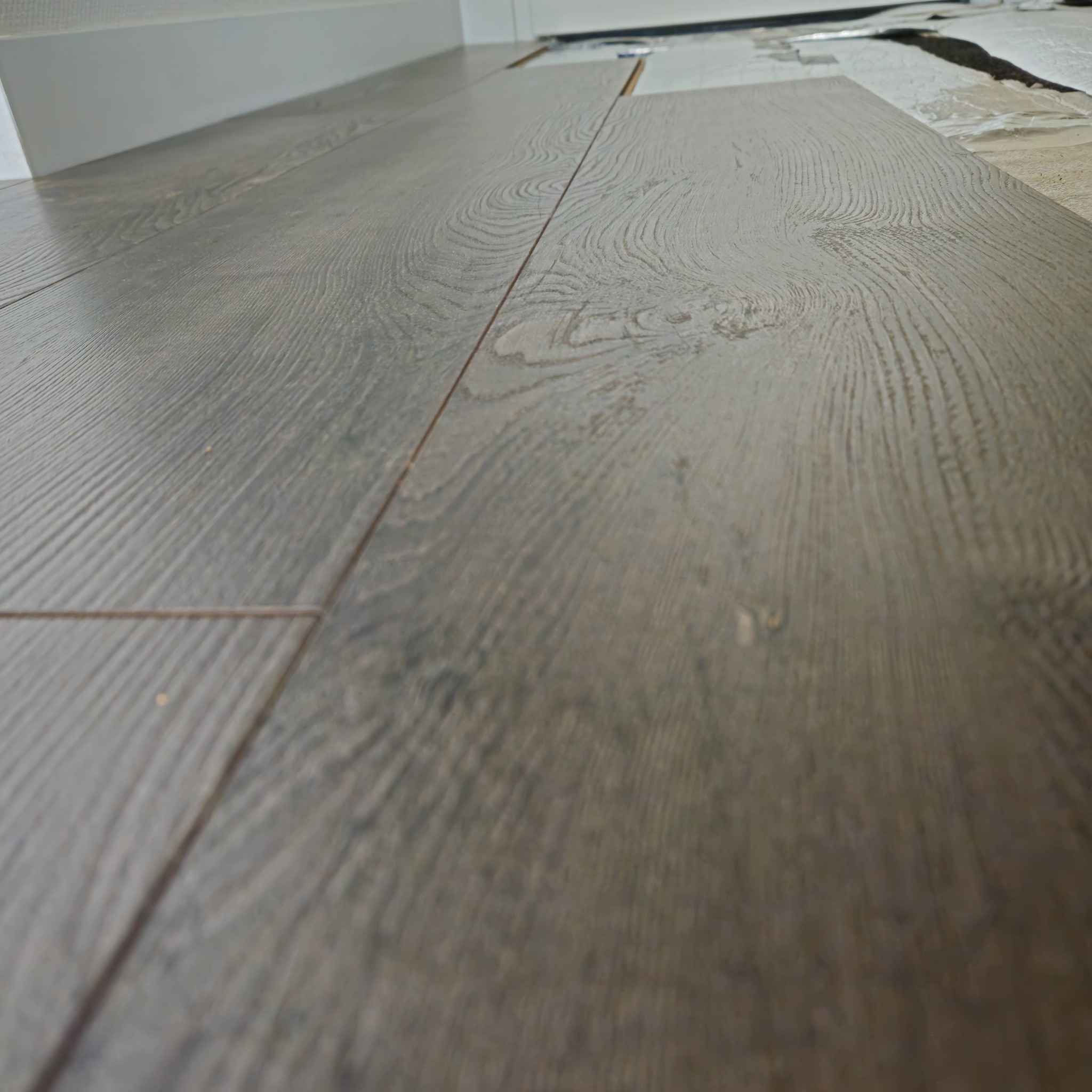 Układanie paneli podłogowych z widoczną strukturą drewna, fragment podkładu i listwa przypodłogowa w tle.