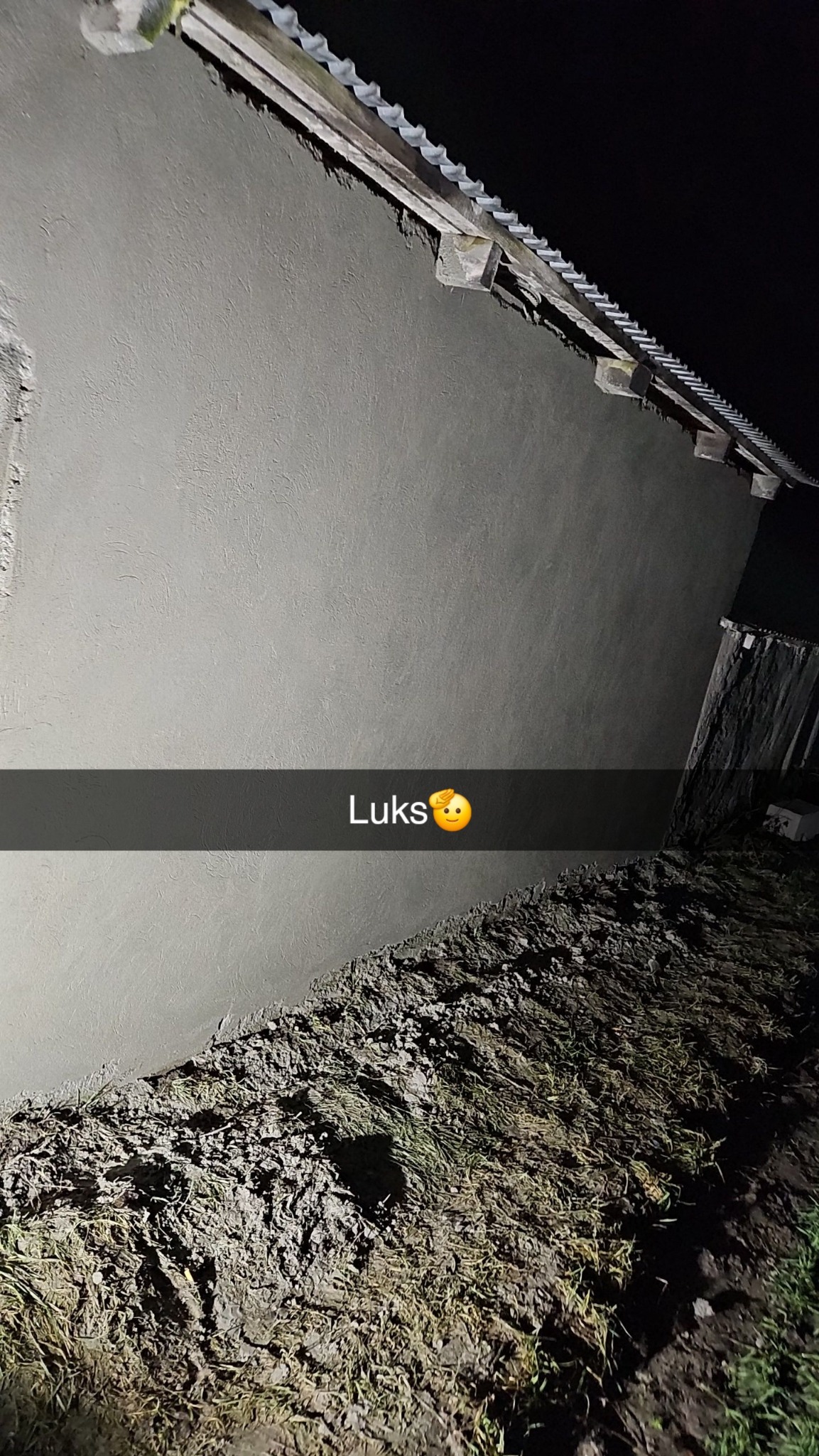 Świeżo otynkowana ściana domu z widoczną strukturą tynku, oświetlona od dołu, z ziemią i fragmentem dachu w kadrze. Na zdjęciu widoczny napis 'Luks' z emotikoną.