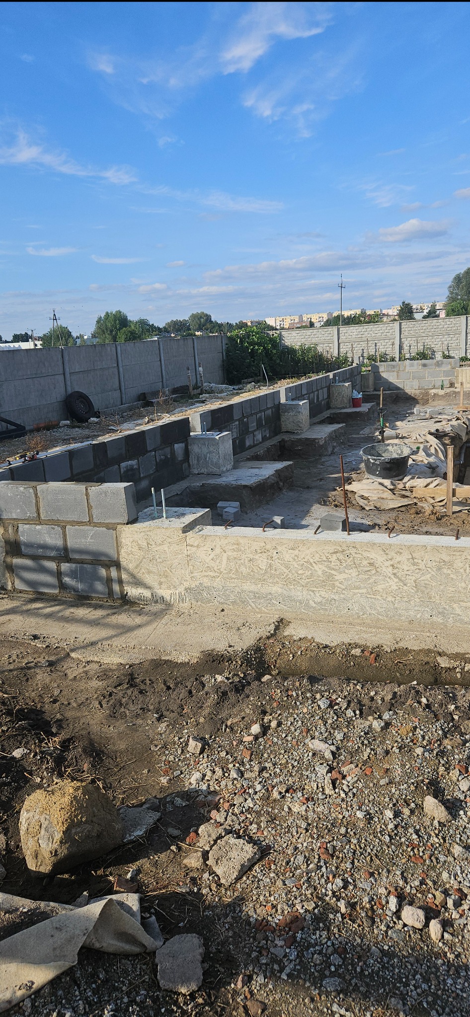 Wykopane fundamenty z częściowo wylanym betonem, widoczne szalunki i bloczki betonowe, w tle ogrodzenie z betonowych płyt i zabudowa miejska pod błękitnym niebem.