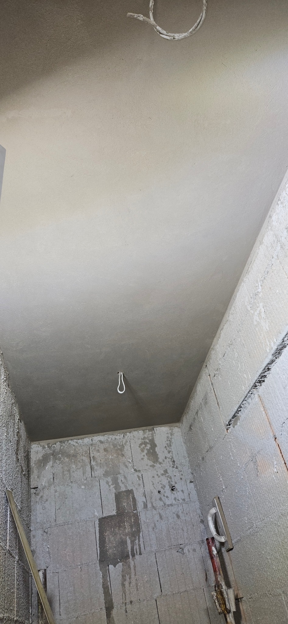 Wnętrze w trakcie prac wykończeniowych: gładki, jasnoszary sufit po szpachlowaniu, surowe ściany z betonu komórkowego z widocznymi śladami zaprawy, wystający kabel oświetleniowy, narzędzia...