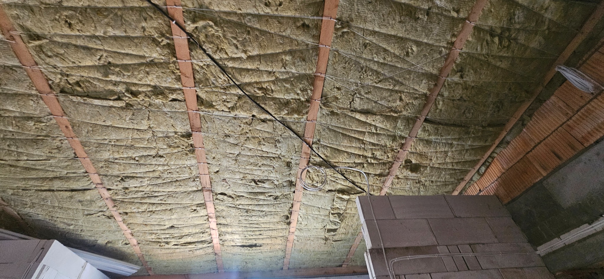 Izolacja poddasza wełną mineralną, widoczne drewniane krokwie i przewody elektryczne, surowy stan wykończenia.