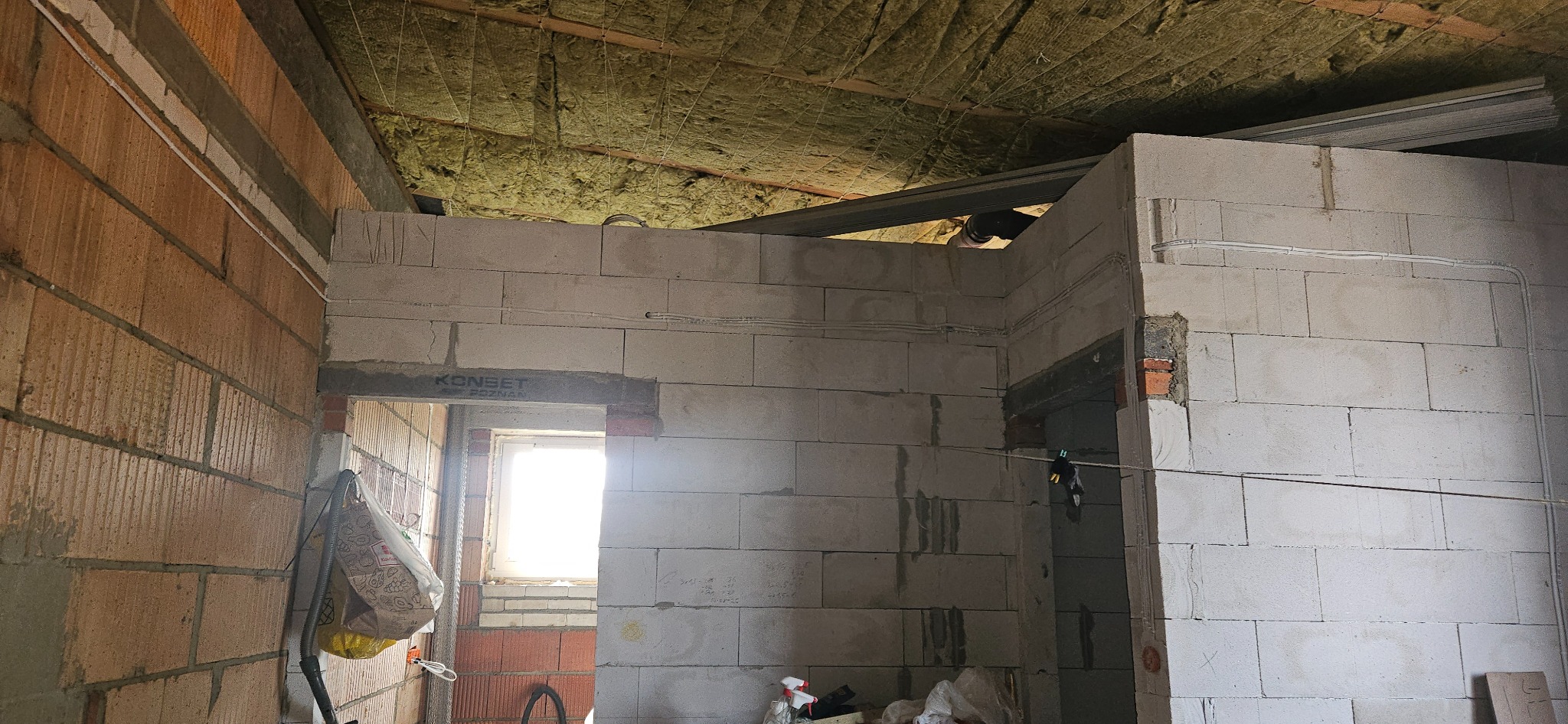 Wnętrze surowego domu w trakcie budowy: ściany z białych bloczków betonowych, częściowo otynkowane, z widocznymi przewodami elektrycznymi, izolacja z wełny mineralnej na suficie, surowa podłoga...