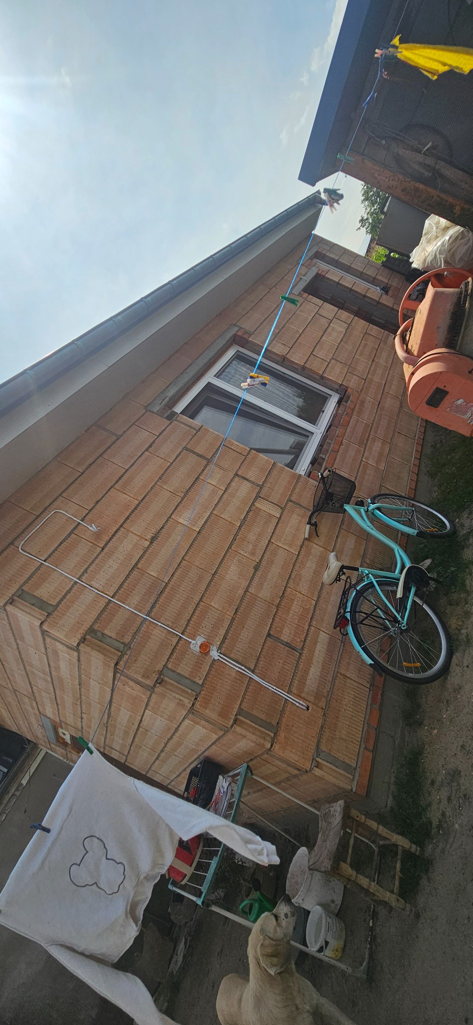 Ściana domu w trakcie budowy z widocznymi pustakami ceramicznymi, okno wstawione, na ścianie poprowadzona instalacja elektryczna, oparty o ścianę rower, w tle betoniarka i rozwieszone pranie.