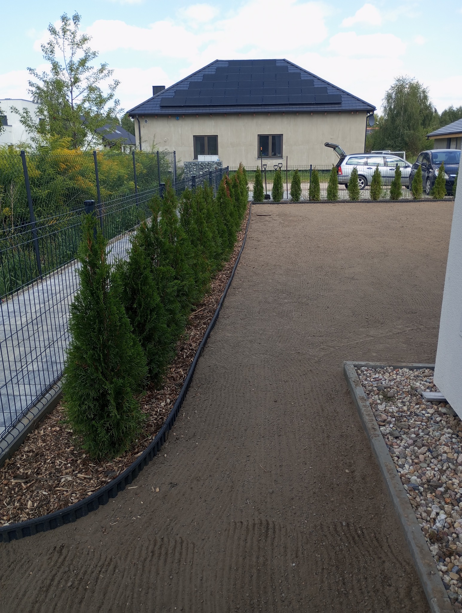 Przygotowany teren pod trawnik z posadzoną linią tuj wzdłuż ogrodzenia, widoczne ślady po bronowaniu, w tle dom z panelami słonecznymi na dachu oraz samochody.