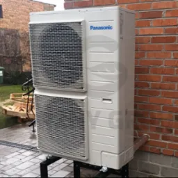 Biała, dwuczęściowa pompa ciepła Panasonic zamontowana na czarnej, metalowej podstawie przy ceglanej ścianie budynku, z widocznym odpływem kondensatu i paletą w tle.