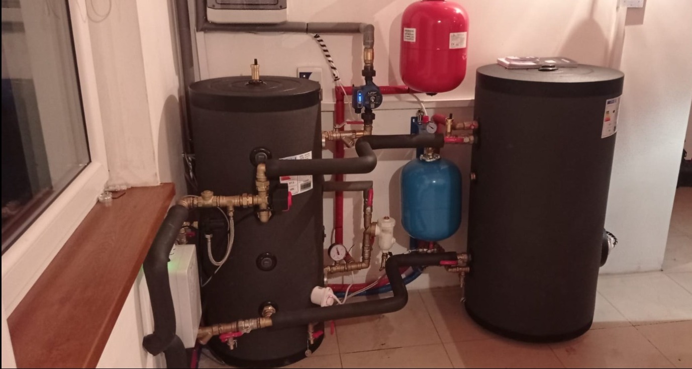 Dwa zbiorniki akumulacyjne ciepła w kotłowni, połączone rurami z zaworami, manometrami i pompą obiegową, widok z boku na tle białych ścian i okna z drewnianym parapetem.