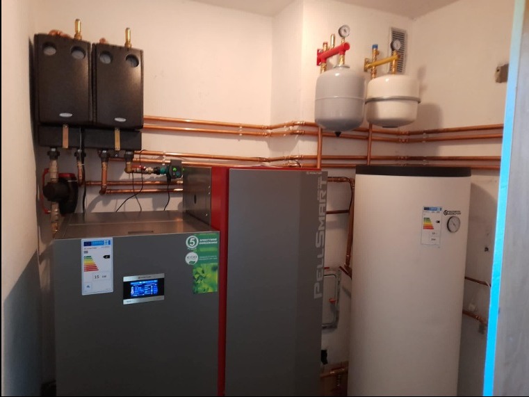 Nowoczesna kotłownia z dwoma piecami, zbiornikiem akumulacyjnym i rozbudowaną instalacją miedzianą, widoczne etykiety energetyczne i manometry.