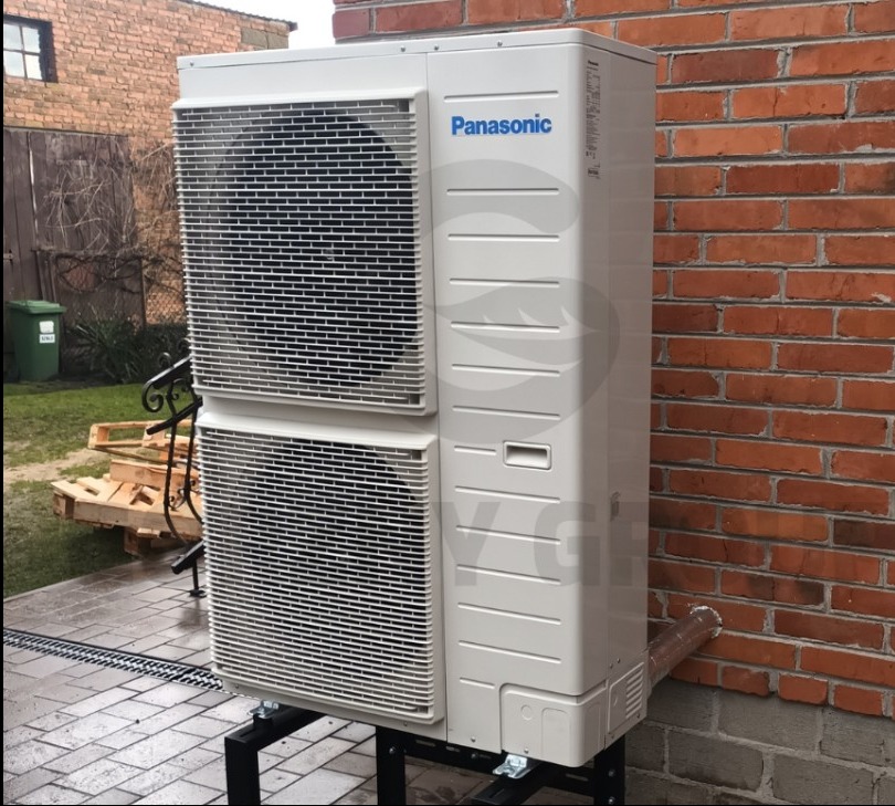 Biała, dwuczęściowa pompa ciepła Panasonic zamontowana na czarnej, metalowej podstawie przy ceglanej ścianie budynku, z widocznym odpływem kondensatu i paletą w tle.