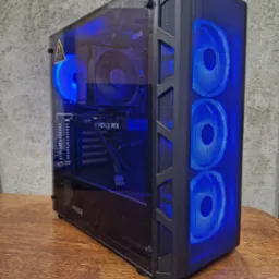 Komputer stacjonarny w czarnej obudowie z przezroczystym panelem bocznym, podświetlony na niebiesko, widoczne komponenty MSI i Gigabyte, stoi na drewnianym blacie na tle szarej ściany.