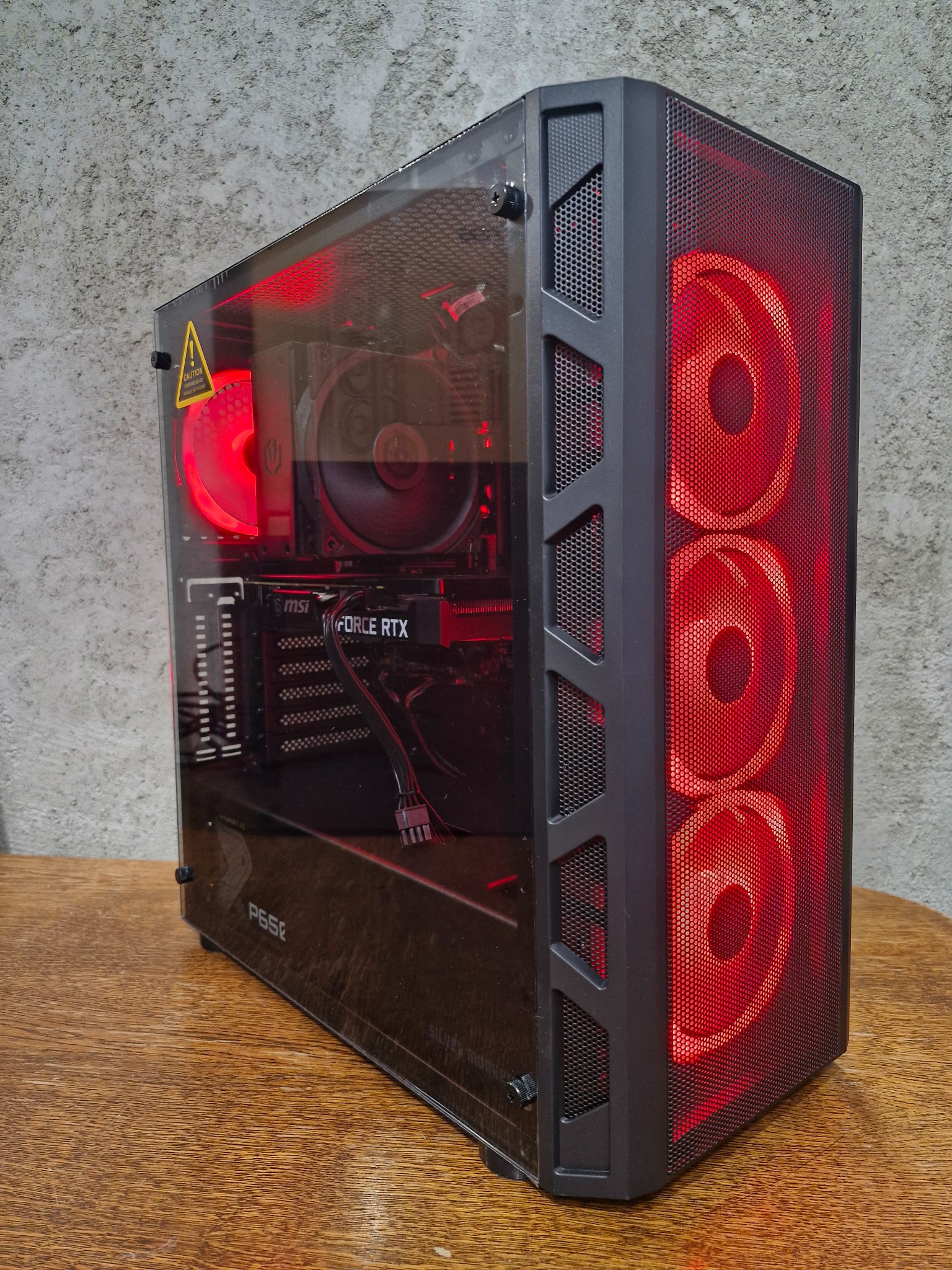 Komputer stacjonarny w czarnej obudowie z przezroczystym panelem bocznym, podświetlony na czerwono z trzema wentylatorami RGB z przodu i widoczną kartą graficzną GeForce RTX wewnątrz, stojący...