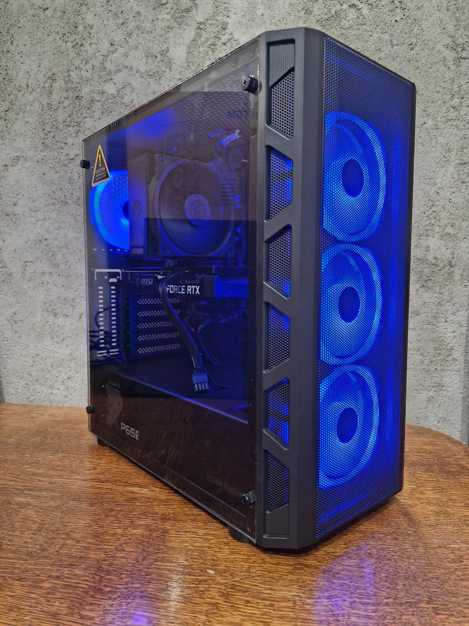 Komputer stacjonarny w czarnej obudowie z przezroczystym panelem bocznym, podświetlony na niebiesko, widoczne komponenty MSI i Gigabyte, stoi na drewnianym blacie na tle szarej ściany.