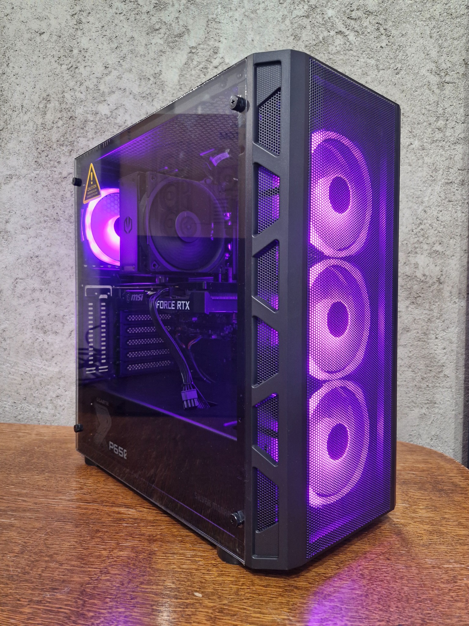 Komputer stacjonarny z podświetleniem LED w obudowie z oknem, widoczna karta graficzna MSI Force RTX, chłodzenie procesora i wentylatory z fioletowym podświetleniem, ostrzeżenie o ryzyku porażenia...