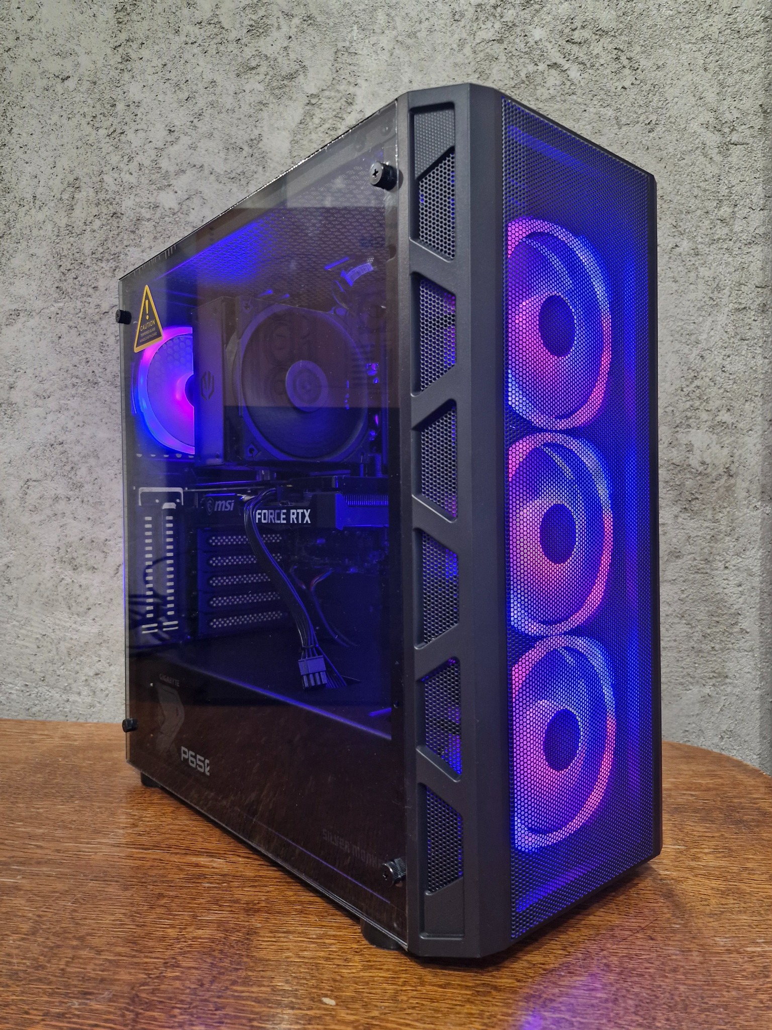 Komputer stacjonarny z przezroczystą obudową, podświetlany wentylatorami RGB, karta graficzna MSI Force RTX, widoczne komponenty na drewnianym blacie.