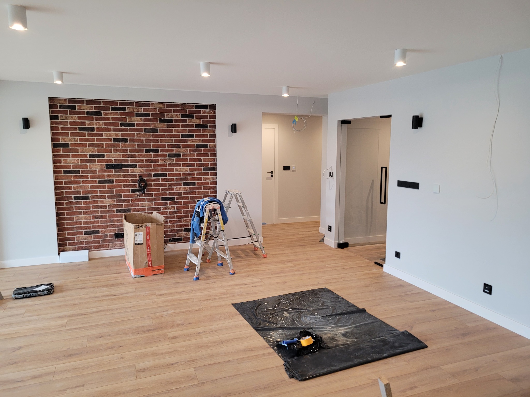 Wnętrze w trakcie montażu oświetlenia: widoczne lampy sufitowe, ściana z cegły dekoracyjnej, drabina malarska z narzędziami, folia ochronna na podłodze i kartonowe pudło.