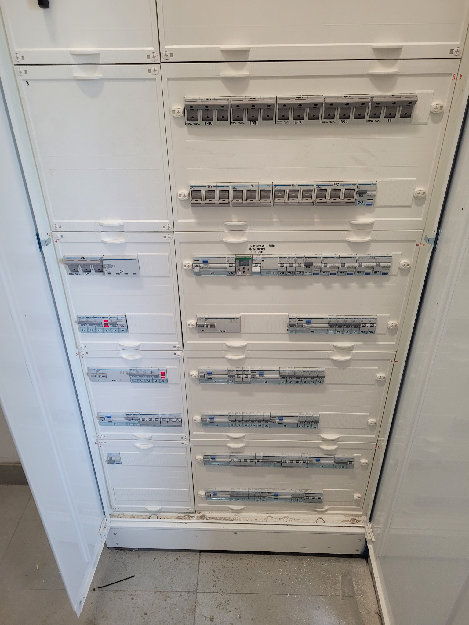 Rozdzielnica główna z widocznymi bezpiecznikami i wyłącznikami różnicowoprądowymi w białej obudowie, etykiety opisujące poszczególne obwody, widok z dołu pod lekkim kątem.