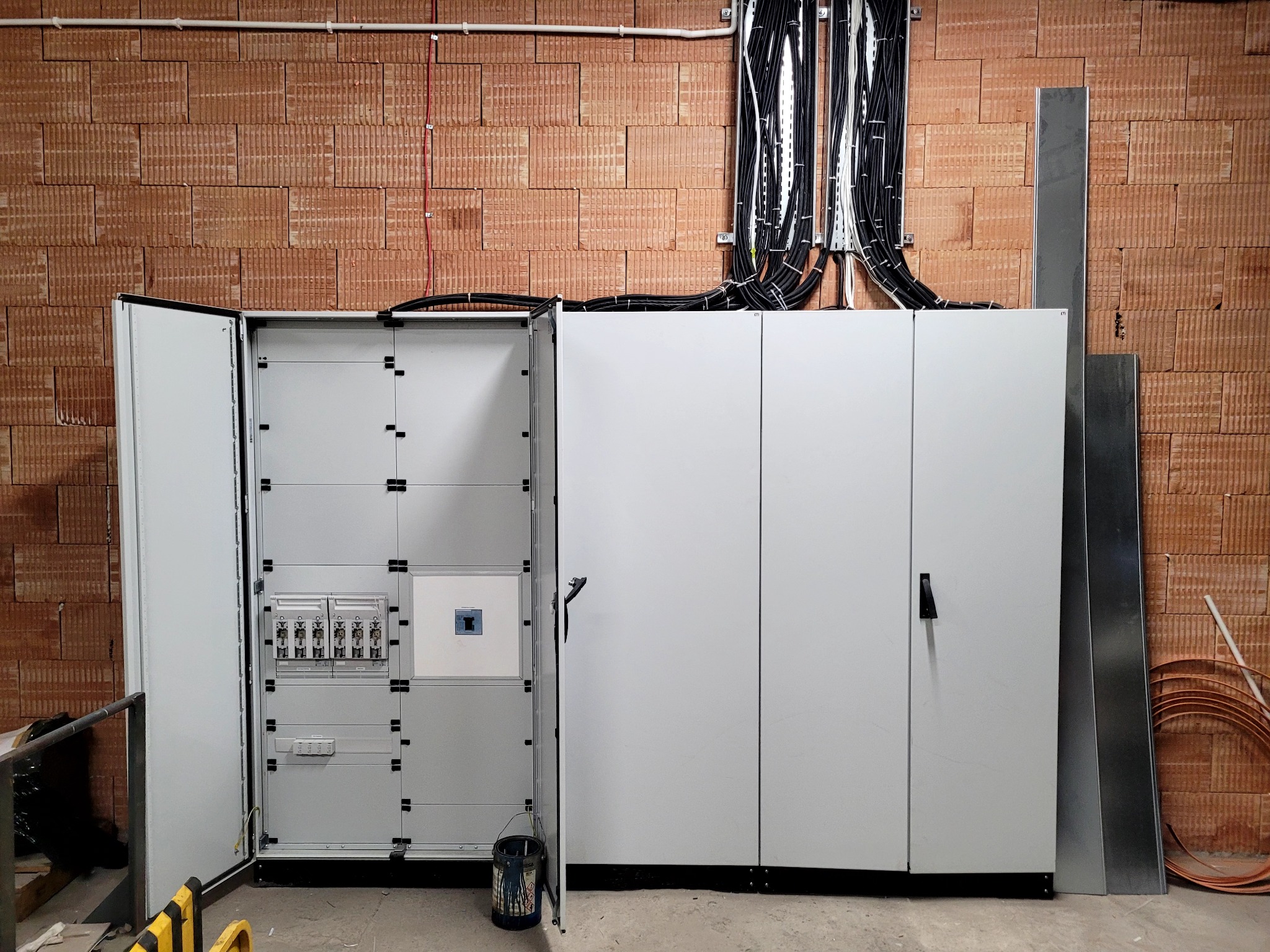 Otwarta rozdzielnica główna z widocznymi bezpiecznikami i okablowaniem, zamontowana na ścianie z cegły, widoczny fragment rur miedzianych i puszka z farbą na podłodze.